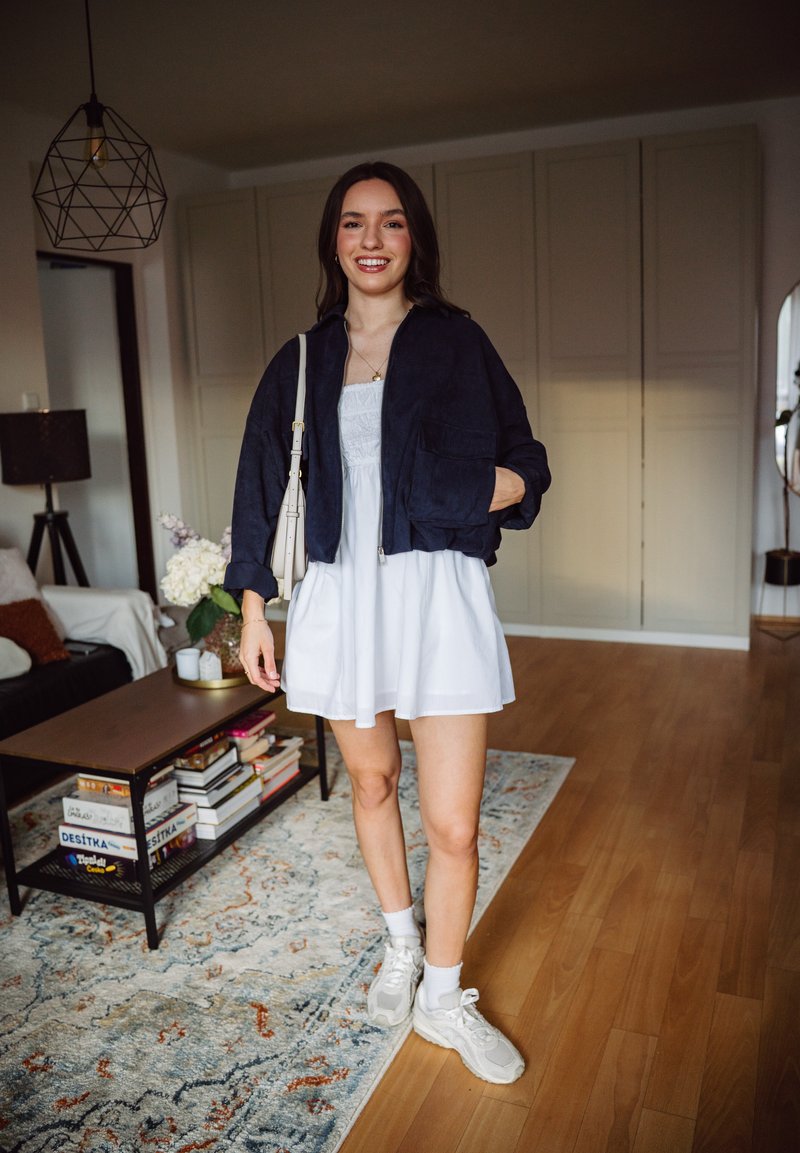 Jeune femme souriante debout dans un salon, portant une robe blanche, une veste noire, des baskets blanches et portant un sac à bandoulière beige.