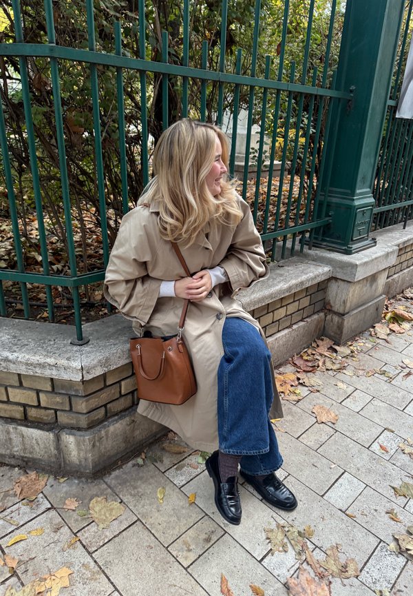 Beige trenchcoat, blauwe wijde jeans, zwarte instappers en een bruin handtas, zittend op een stenen bank met herfstbladeren. Groene hekken op de achtergrond.