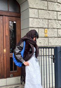 Sinine tikandiga kott, pruun tekstuuriga sall, nahktagi, hall top ja valge mustriline seelik. Seisab puidust ukse ees kiviseina taustal.