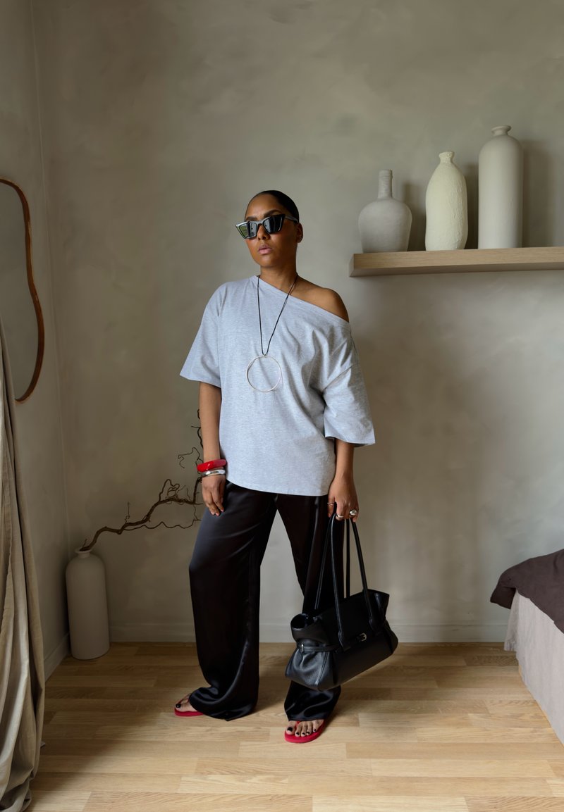 Femme portant un top gris oversize à épaules dénudées, pantalon noir à jambes larges, tongs rouges, sac à main noir, lunettes de soleil et grand collier circulaire à l'intérieur.