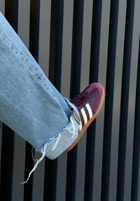 Basket de sport bordeaux et crème en daim texturé, avec trois bandes blanches et une semelle en caoutchouc gum. Un jean bleu clair effiloché laisse apparaître la cheville.