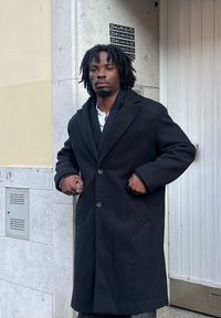 Czarny wełniany płaszcz z klapowym kołnierzem, dużymi guzikami i bocznymi kieszeniami. Noszony na białej koszuli, prezentowany na zewnątrz.