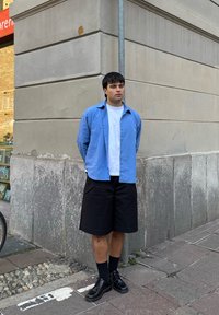 Camisa azul claro abotonada sobre una camiseta blanca, combinada con pantalones cortos negros y zapatos negros brillantes; de pie contra una pared de piedra.