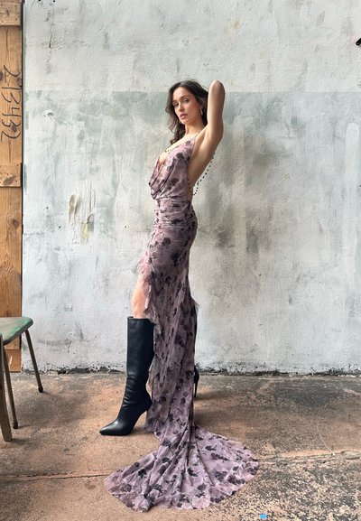 Vestido floral morado con diseño drapeado, abertura y falda fluida. Un par de altas botas de tacón de aguja negras realza el look. Textura de tela suave.