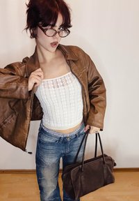 Veste en cuir marron portée sur un crop top en dentelle blanche, assortie à un jean bleu délavé. Un sac à main sombre est tenu dans une main. Fond simple.