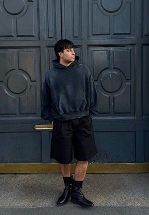 Joven con sudadera oscura de gran tamaño, pantalones cortos negros y botas negras, de pie con las manos en los bolsillos frente a grandes puertas dobles azules.