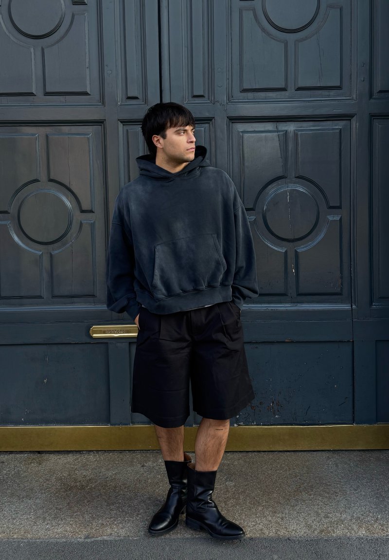Jeune homme portant un sweat à capuche sombre et oversized, des shorts noirs et des bottes noires, debout les mains dans les poches devant de grandes portes doubles bleues.