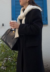 Γυναίκα με μαύρο παλτό και λευκό κασκόλ κρατά μαύρη τσάντα σε ένα χέρι και μια κούπα καφέ στο άλλο, κοντά σε μπλε παντζούρια.