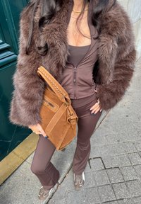 Braune Kunstfelljacke, dunkelbrauner Reißverschlussoberteil, passende Schlaghosen und eine hellbraune Wildlederhandtasche mit geflochtenen Details und Hardware-Akzenten.