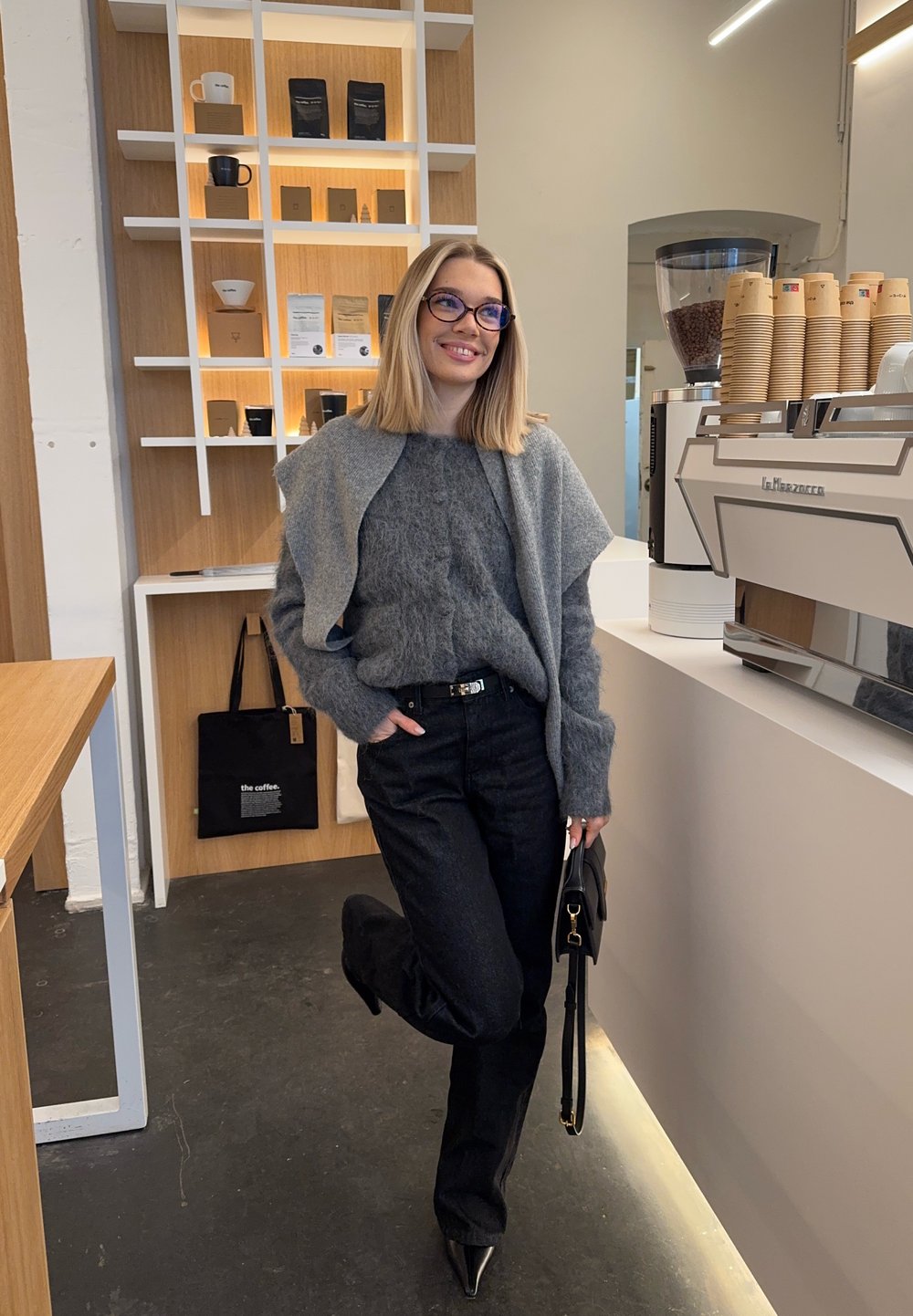 Femme portant des lunettes, un pull gris et un pantalon noir, debout près d'une machine à café dans un café moderne, tenant un sac à main noir.