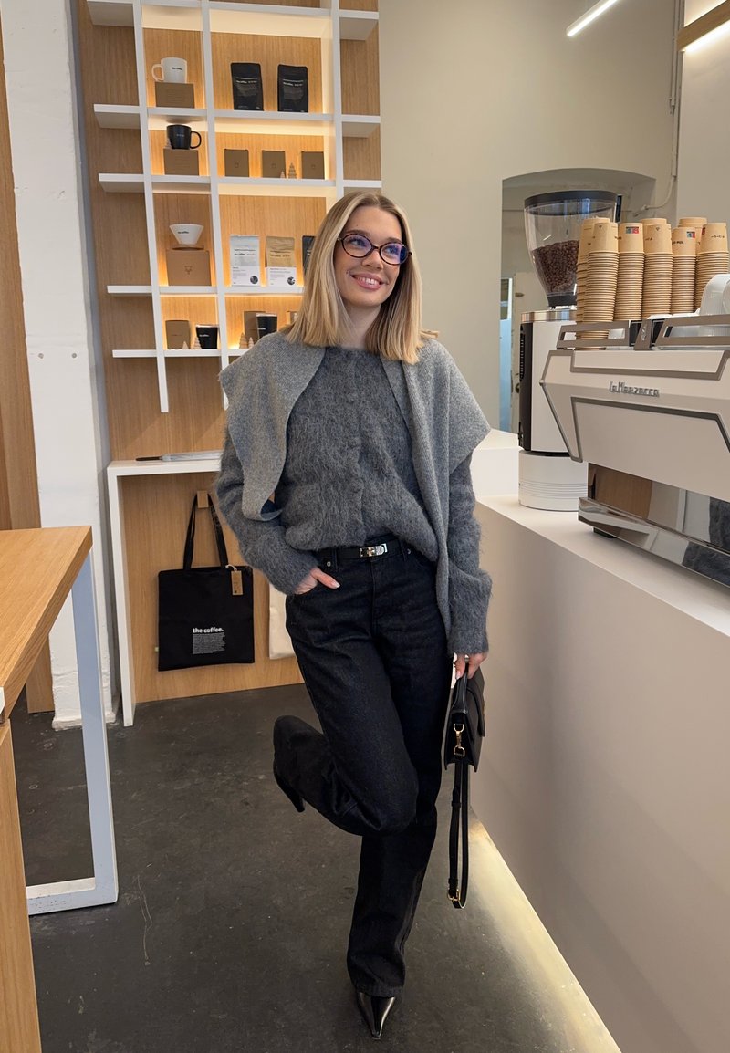 Femme portant des lunettes, un pull gris et un pantalon noir, debout près d'une machine à café dans un café moderne, tenant un sac à main noir.