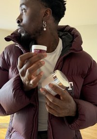 Homme en veste bordeaux tenant deux pots de soins pour la peau, regardant sur le côté, avec des boucles d'oreilles et des tatouages sur le cou visibles.