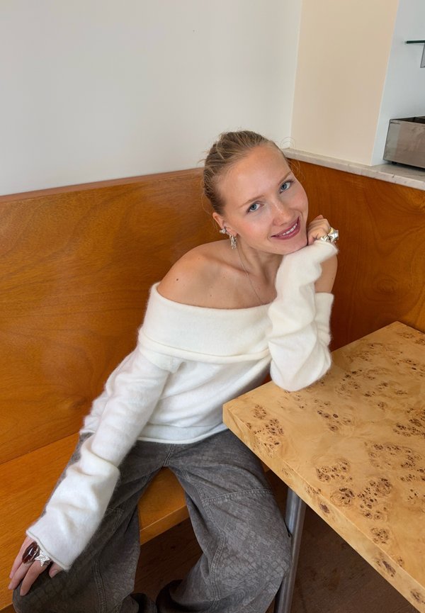 Pull en tricot blanc décolleté, pantalon gris à jambes larges avec motif, bague et boucles d'oreilles. Table en bois et marbre dans un éclairage clair.