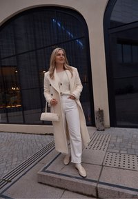 Casaco bege, blusa de malha branca, calças brancas de cintura alta e loafers a condizer. Pequena bolsa em bege claro. Fundo arquitetónico.