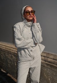 Outfit bianco strutturato con colletto alto, bottoni a pressione e tessuto con motivi. Accompagnato da occhiali da sole marroni oversized.