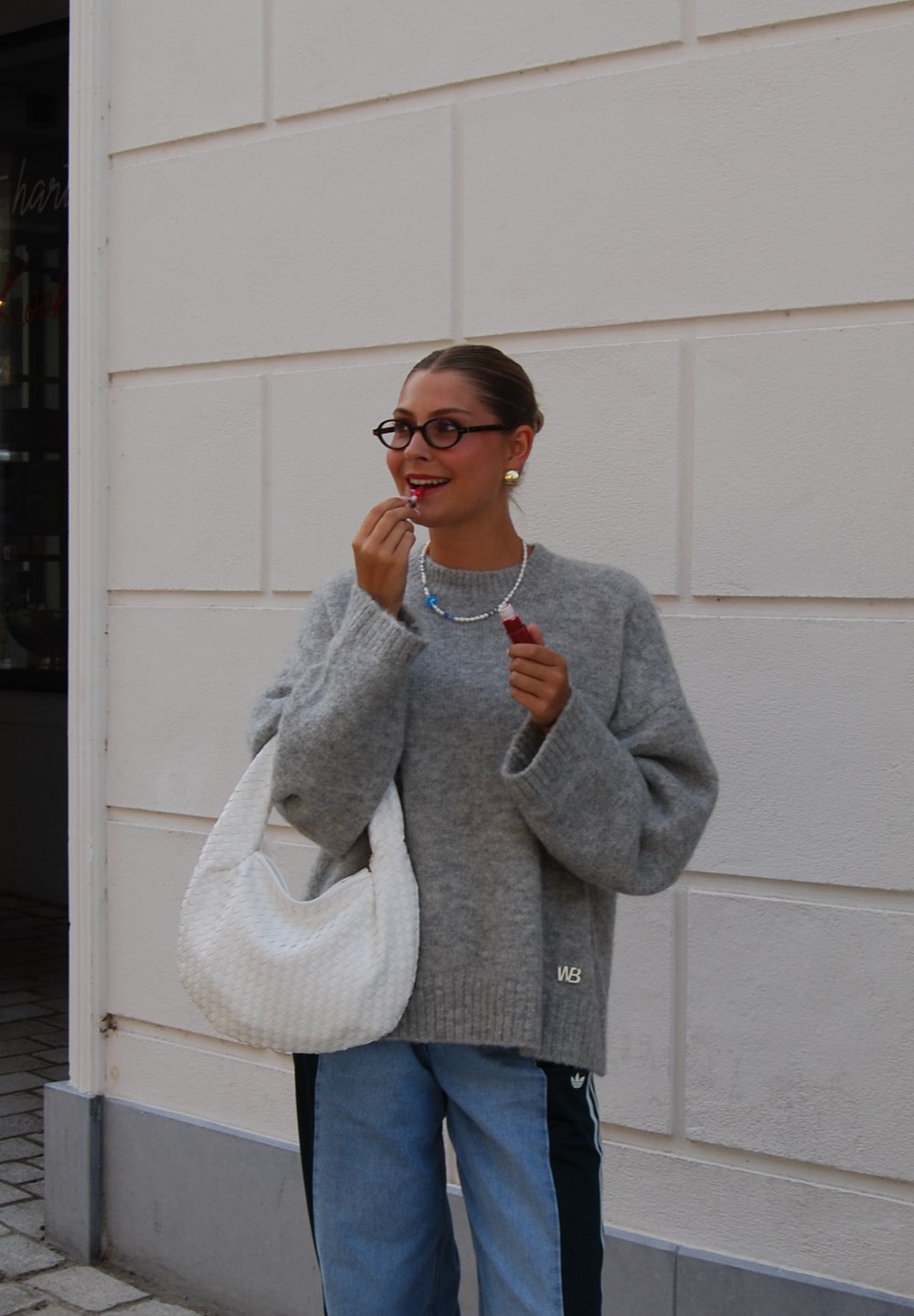 Grå oversized genser, lyseblå jeans, hvit vevd håndveske, fargerik halskjede, runde briller, påfører leppestift. Utendørs setting.