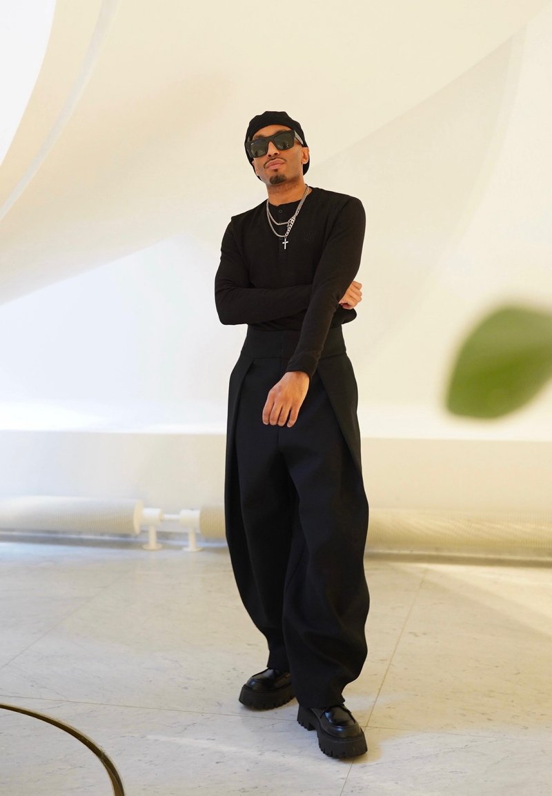 Homme portant une chemise noire à manches longues, un pantalon noir oversize, une casquette noire, des lunettes de soleil et des chaussures épaisses, debout les bras croisés à l'intérieur.