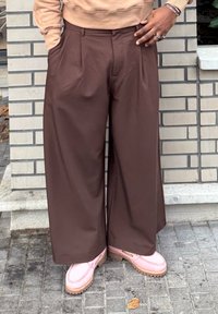 Pantalon marron à jambes larges avec plis, affichant une texture lisse. Associé à des chaussures roses sans lacets avec une semelle texturée.