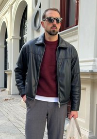 Czarna skórzana kurtka z zamkiem z przodu, na burgundowym swetrze. Szare spodnie, trzymające beżową torbę. Gładkie okulary przeciws�łoneczne dopełniają stylizację.