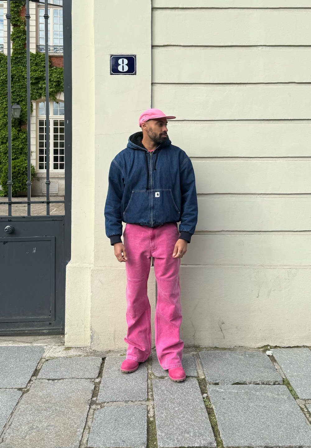 Τζάκετ από τζιν με κουκούλα, μαύρη εσωτερική επένδυση, ροζ φαρδιές τζιν, αντίστοιχο ροζ σκουφί και παπούτσια; στέκεται απέναντι σε έναν ανοιχτόχρωμο τοίχο με πράσινα φυτά.