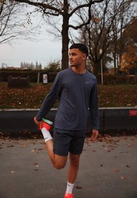 Tenue sportive comprenant un haut à manches longues bleu marine, un short gris, et des baskets rose vif avec des semelles vertes. Le cadre comprend des feuilles tombées.