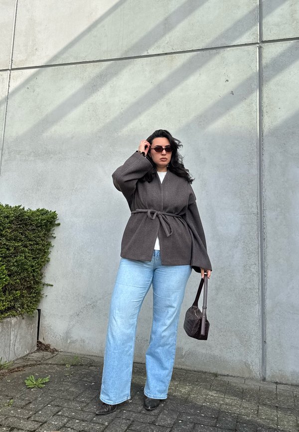 Femme aux cheveux foncés portant des lunettes de soleil, un manteau gris ceinturé, un jean bleu clair, des chaussures noires, tenant un sac à main foncé, debout contre un mur en béton.