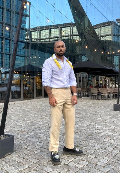 Hombre vestido con una camisa de botones a rayas azul claro, pantalones beige y zapatos negros con acentos gráficos, de pie sobre una superficie de adoquines.