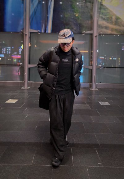 Chaqueta acolchada negra, cuello alto negro, pantalones anchos a rayas y gorra de béisbol. Mochila negra y zapatillas. Fondo urbano con poca iluminación.