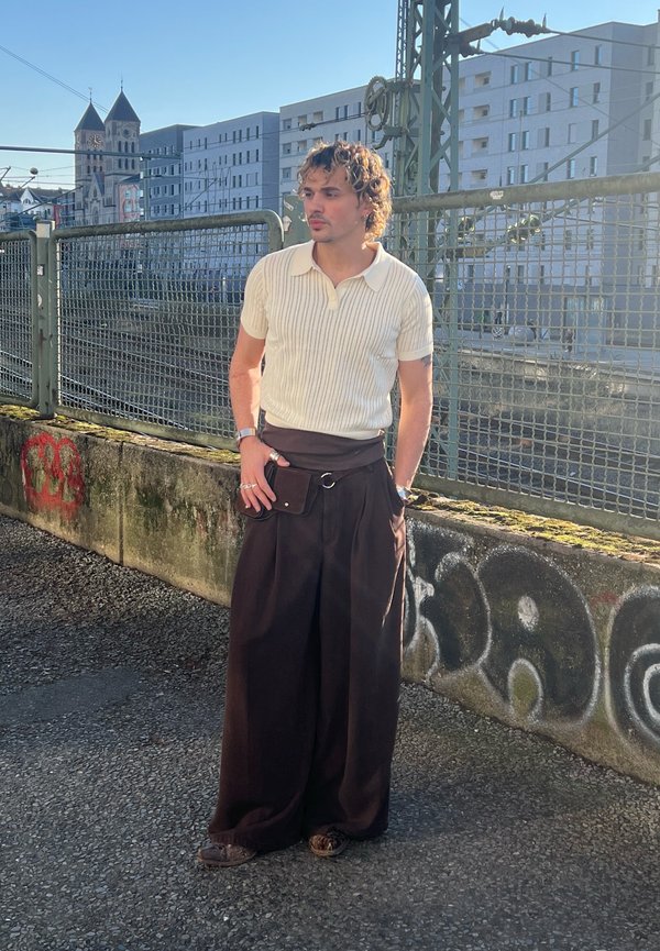 Noor mees lokkis juustega, valges lühikeste varrukatega kootud särgis ja laiade mustade pükstega, seisab grafitidega kaetud betoonseina kõrval metallaia juures.