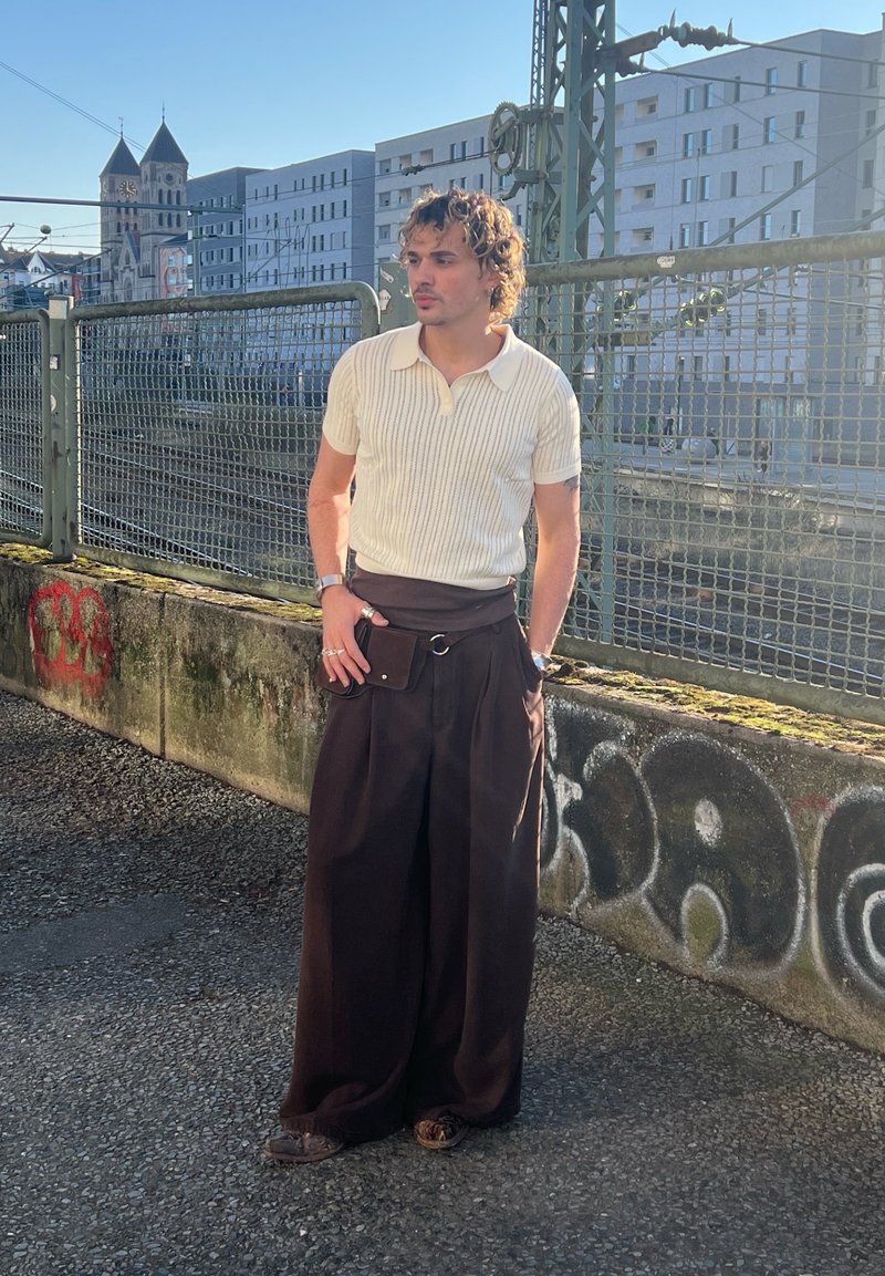 Jeune homme aux cheveux bouclés portant un t-shirt en maille à manches courtes blanc et un large pantalon noir, debout près d'un mur en béton couvert de graffitis, près d'une clôture en métal.