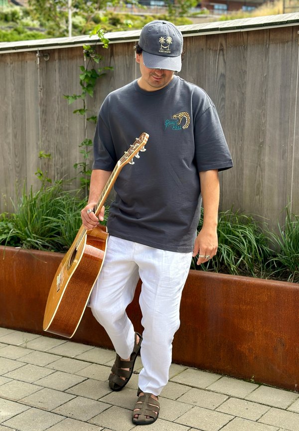 Hombre con camiseta oscura y gorra sostiene una guitarra acústica mientras está de pie en un camino pavimentado junto a una cerca de madera y vegetación.