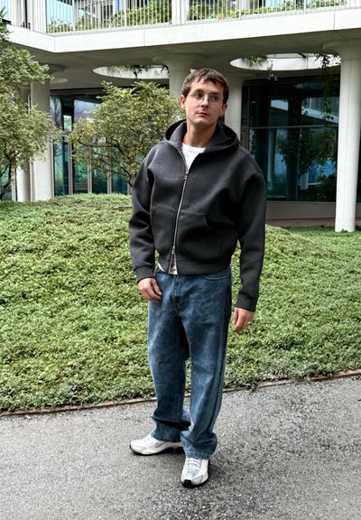 Sudadera gris con cremallera y capucha grande, llevada sobre una camiseta blanca, combinada con vaqueros vaqueros azul claro. Zapatillas deportivas cómodas completan el conjunto.