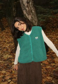 Turkusowa polarowa kamizelka z guzikiem z przodu i granatowym wykończeniem, noszona na białej koszuli z długim rękawem; na tle jesiennego krajobrazu z opadłymi liśćmi.