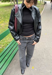 Persona, kas valkā melnu ādas jaku, melnu augstu apkakli, pelēnas džinsas, melnas zābaki un rokās tur bordo krāsas pinumu somu, stāv pie zaļas parka sola.