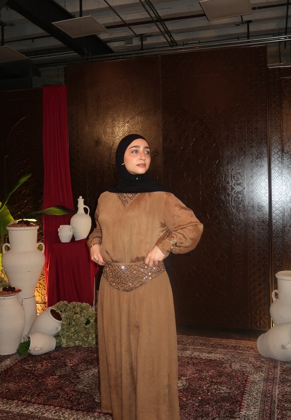 Mujer con vestido marrón y hiyab negro de pie en una habitación ornamentada con grandes jarrones blancos y una alfombra con diseño.