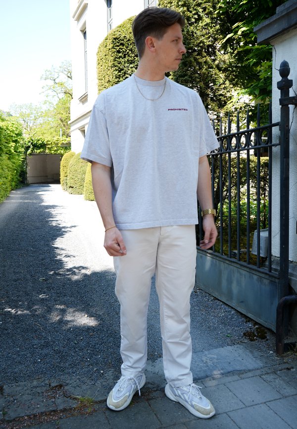 Camiseta de manga corta gris con texto rojo, combinada con pantalones cargo blancos y zapatillas ligeras. Entorno al aire libre con vegetación y una puerta.