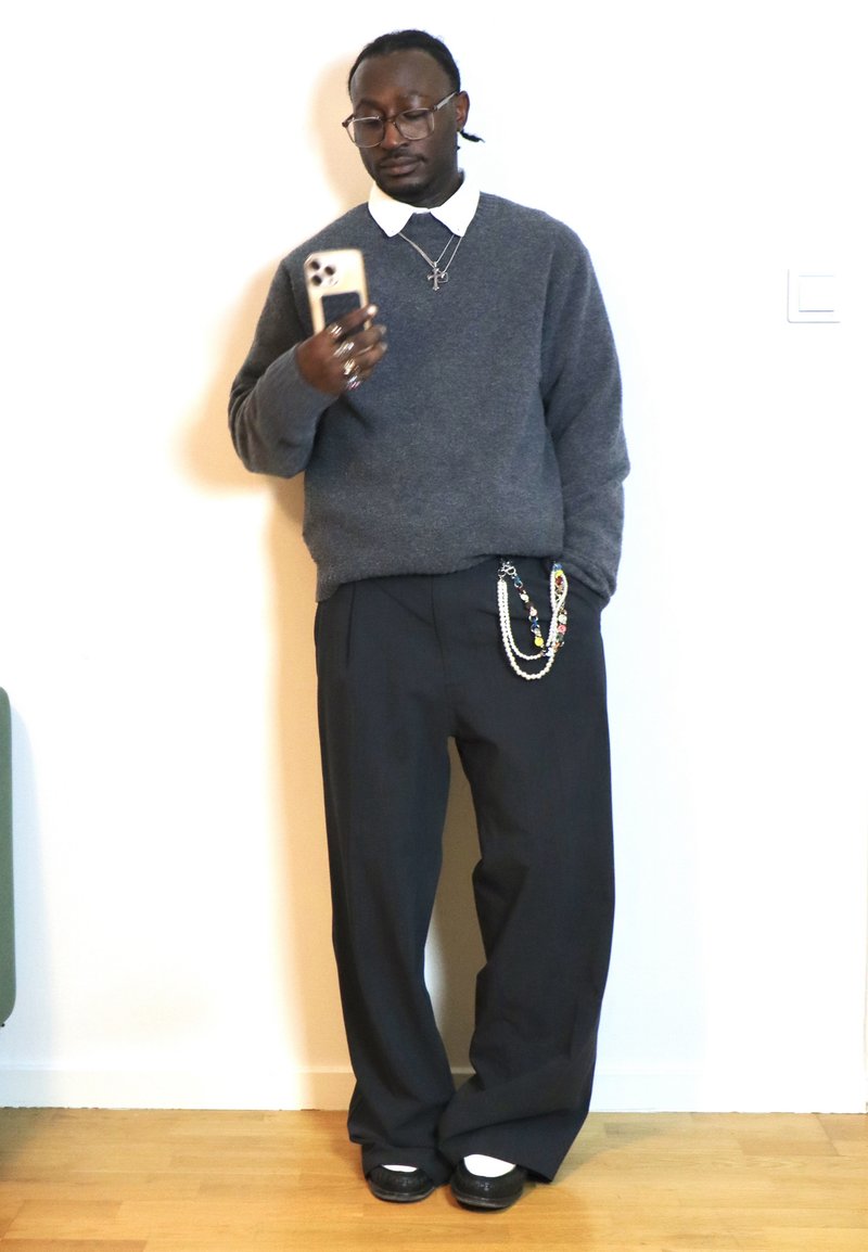 Man wearing grey jumper, white collared shirt, black wide trousers with decorative chains, black shoes, holding smartphone while standing indoors.