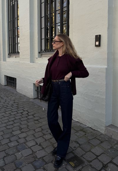 Blazer burdeos oscuro sobre una blusa ajustada burdeos, combinado con unos vaqueros de tiro alto azul oscuro. Un bolso negro acolchado y zapatos negros completan el look.