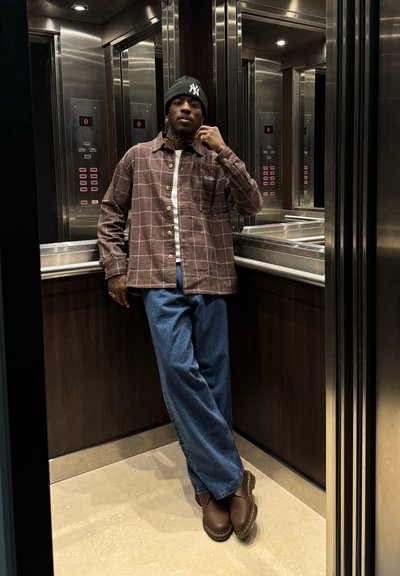 Camisa de franela marrón a cuadros sobre una camiseta blanca, combinada con jeans azules y zapatos marrones. De pie en un ascensor con acabados de metal y madera.