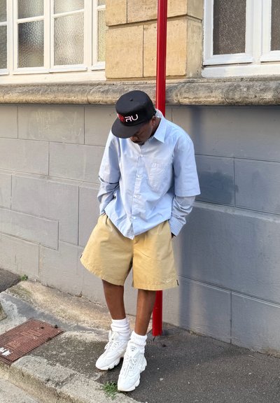 Camisa de botones azul claro con mangas a rayas, pantalones cortos beige, zapatillas blancas y una gorra negra con detalles rojos. Reclinándose contra una pared.