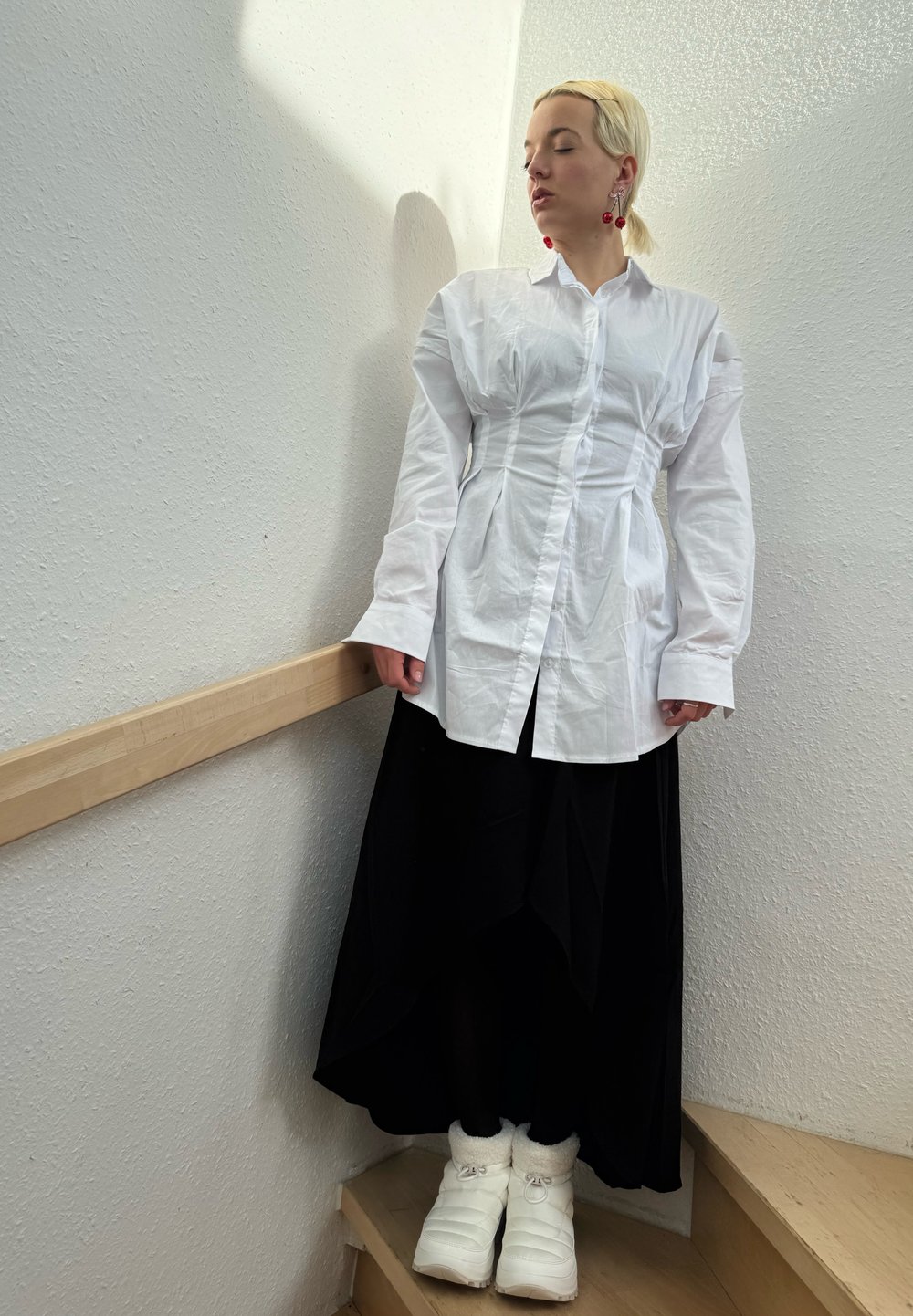 Camisa blanca de botones con cintura fruncida, falda negra larga y botas de invierno blancas. Escalera de madera y pared blanca texturizada al fondo.