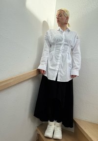 Camisa blanca de botones con cintura fruncida, falda negra larga y botas de invierno blancas. Escalera de madera y pared blanca texturizada al fondo.
