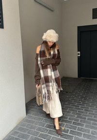 Brown oversized sweater, plaid scarf featuring shades of brown and white, beige skirt, brown heeled shoes, beige bag, and a white fur hat. Background of brick pavement.