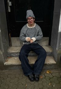 Preveliki prugasti pulover u tamnoplavoj i krem boji, labave traperice, crne čipkaste čizme, sivi beanie; sjedi na stepenicama s teksturiranom površinom.
