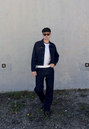 Hombre con gafas de sol, gorra negra, chaqueta vaquera y vaqueros, de pie sobre terreno rocoso frente a una pared gris agrietada con señales numeradas 4 y 5.