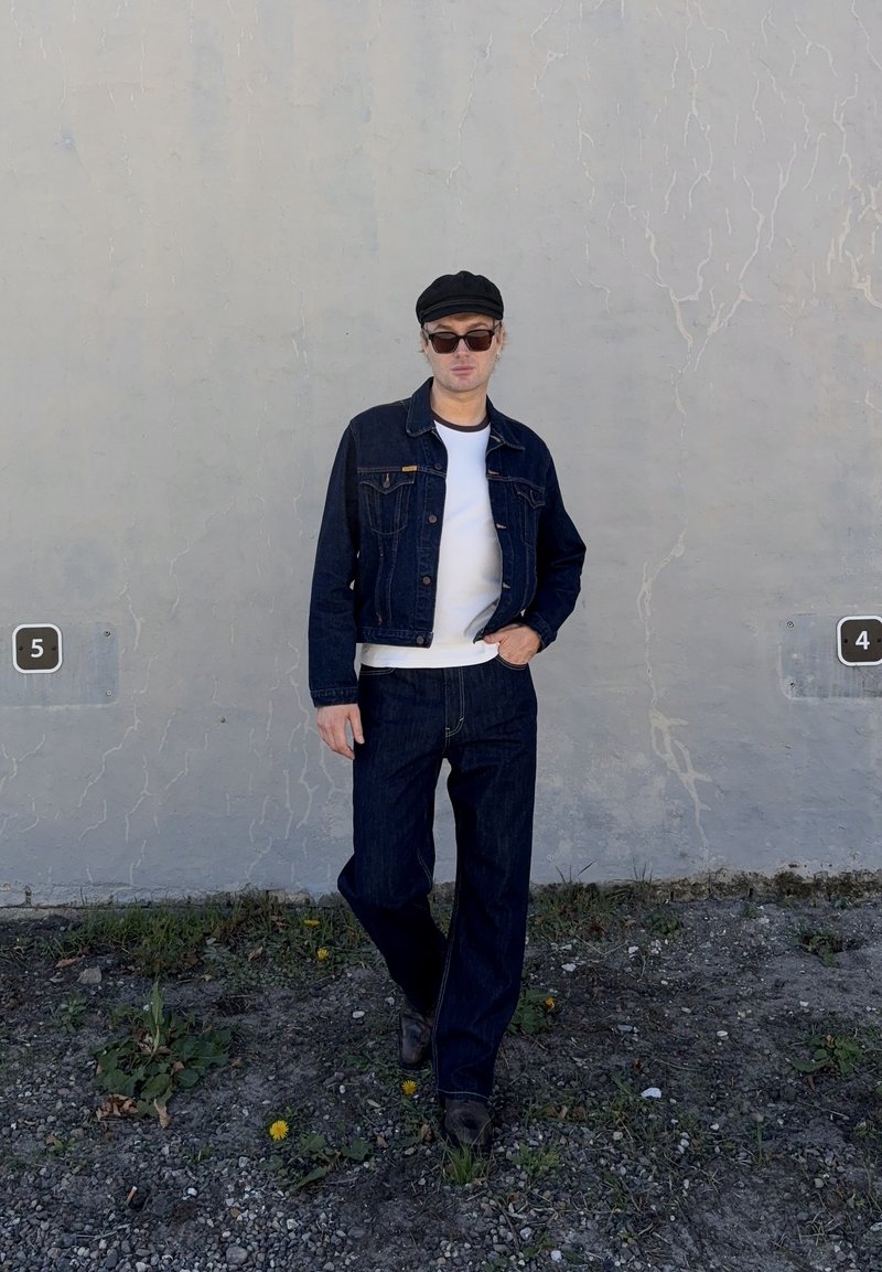 Homme portant des lunettes de soleil, une casquette noire, une veste en jean et un jean, debout sur un sol rocheux contre un mur gris fissuré avec des panneaux numérotés 4 et 5.