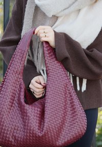 Μπορντώ πλεκτή δερμάτινη τσάντα με μαλακή, στρογγυλεμένη φόρμα και υφή, κρατημένη από ένα άτομο που φοράει καφέ πουλόβερ και γκρι scarf.