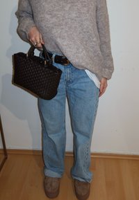 Personne portant un pull beige, un jean bleu clair retroussé, des chaussures en daim marron, tenant un sac à main tissé foncé par les poignées.