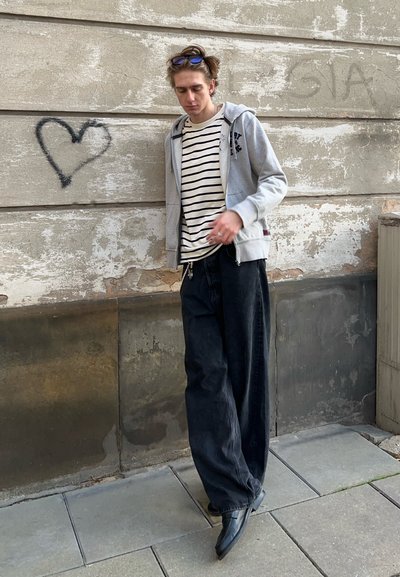 Sudadera gris con cremallera sobre una camiseta de rayas blanca y negra, combinada con pantalones holgados negros y zapatos negros de punta, contra una pared texturizada.