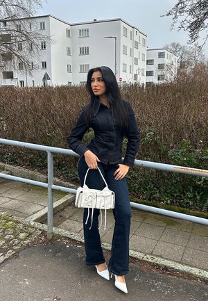 Chaqueta de mezclilla negra, jeans de campana azul oscuro, tacones blancos de punta y un pequeño bolso blanco con detalles de cordón; ambiente urbano al aire libre.